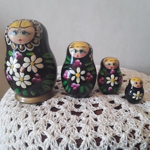 May include: A set of four black nesting dolls featuring hand-painted yellow hair, blue eyes, and red lips. Each doll is decorated with white daisy flowers, purple accents, and green leaves. The largest doll has a gold-toned base at the bottom.