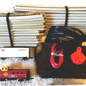 May include: A model train set including gray plastic tracks bundled with black ties, a black power pack with a red control knob, red and black electrical wires, a white Lehigh Valley freight car, a red caboose, and a white Southern freight car.