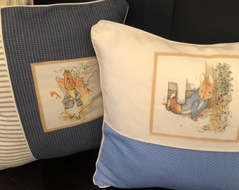 May include: Two decorative throw pillows with blue and white fabric. One pillow features a blue and white striped pattern with a small square image of Peter Rabbit in a garden. The other pillow has a white background with a blue and white polka dot pattern on the bottom half. The image on this pillow shows Peter Rabbit hiding behind a tree.