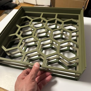 Puede incluir: Panel decorativo cuadrado verde oliva con un patrón de panal. El panel es sostenido por una mano, mostrando el diseño geométrico. El panel está hecho de un material sólido y tiene un efecto tridimensional.