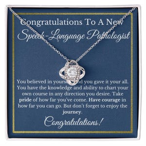 May include: A silver-toned necklace with a pendant featuring a central clear stone surrounded by a swirling design. The necklace is presented in a box with a blue background and the text "Congratulations To A New Speech-Language Pathologist."
