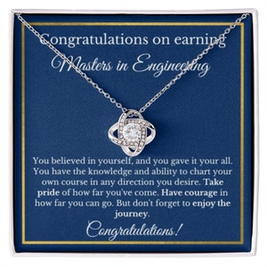 Puede incluir: Un collar plateado con un colgante brillante, presentado en una caja de regalo. La caja tiene un fondo azul marino con el texto "Felicitaciones por obtener una Maestría en Ingeniería". El colgante es un diseño de nudo con una piedra central clara.