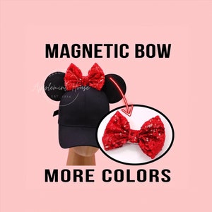May include: A black baseball cap with red sequin bow and mouse ears. The text "MAGNETIC BOW" is at the top of the image. The text "MORE COLORS" is at the bottom of the image.