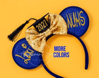 May include: A blue glitter headband featuring two round ears with gold cursive text, a gold sequin bow, and a black miniature graduation cap ornament with the year 2027 printed in gold. The headband has a dark blue fabric-covered band.