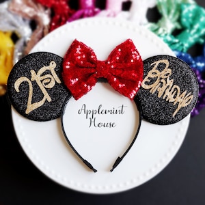 Może przedstawiać: Czarna opaska na włosy z uszami Minnie Mouse z czerwoną kokardą z cekin&oacute;w. Lewy uszek ma złote literki z brokatem, kt&oacute;re m&oacute;wią "21st", a prawy uszek ma złote literki z brokatem, kt&oacute;re m&oacute;wią "Birthday".
