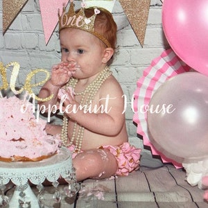 May include: A baby girl wearing a pink and gold glitter crown with the word "One" on it, is sitting on a white wood floor with a pink and white cake in front of her. She is wearing a pink and gold ruffled diaper cover and a pearl necklace. There are pink and gold balloons in the background.