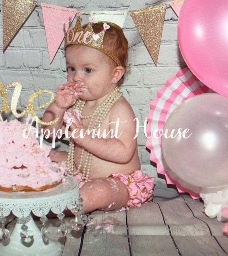 May include: A baby girl wearing a pink and gold glitter crown with the word "One" on it, is sitting on a white wood floor with a pink and white cake in front of her. She is wearing a pink and gold ruffled diaper cover and a pearl necklace. There are pink and gold balloons in the background.