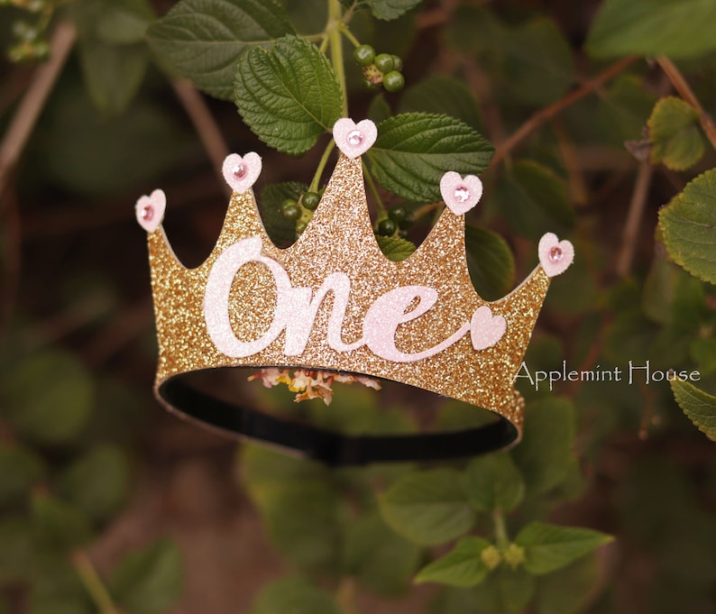 May include: Gold glitter crown headband with pink hearts and the word "One" in pink letters.