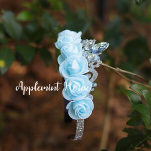 May include: A light blue headband with a silver glitter crown and five light blue fabric roses. The headband has the text "Applemint House" on it.