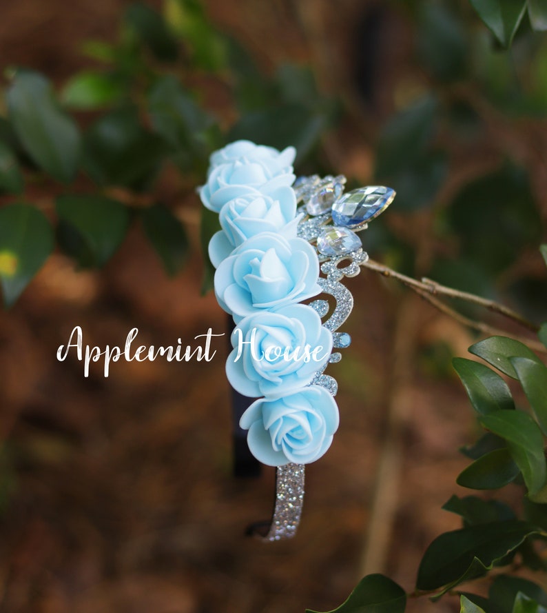 May include: A light blue headband with a silver glitter crown and five light blue fabric roses. The headband has the text "Applemint House" on it.