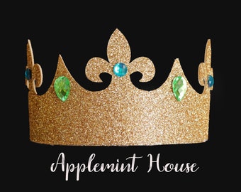 May include: A gold glitter crown featuring a central fleur-de-lis shape, accented with round blue and teardrop-shaped green rhinestones. The text "Applemint House" is printed in white script below the crown against a solid black background.