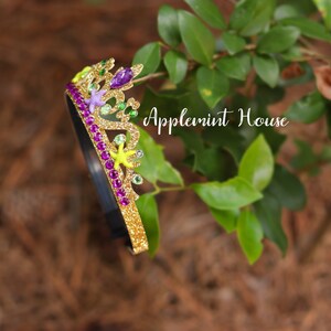 May include: A gold glitter headband with a crown design. The crown is decorated with purple, green, and yellow glitter accents, as well as small plastic sea creatures. The headband is on a green leafy background. "Applemint House" is written in white text on the background.