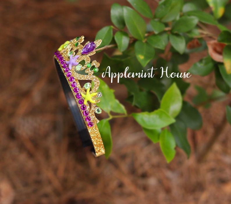 May include: A gold glitter headband with a crown design. The crown is decorated with purple, green, and yellow glitter accents, as well as small plastic sea creatures. The headband is on a green leafy background. "Applemint House" is written in white text on the background.