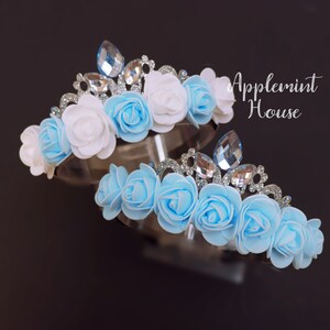 May include: Two flower crowns with blue and white fabric roses and silver glitter accents. The crowns have clear rhinestone embellishments.