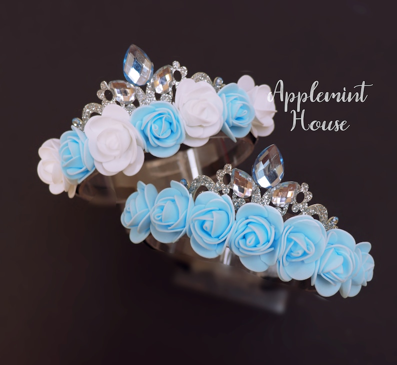 May include: Two flower crowns with blue and white fabric roses and silver glitter accents. The crowns have clear rhinestone embellishments.
