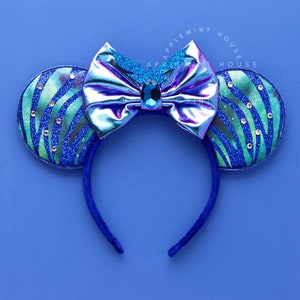 Op de afbeelding: Blauw en groen glitter muis oren haarband met een holografische regenboogkleurige strik en een blauwe strass in het midden. De haarband is blauw.