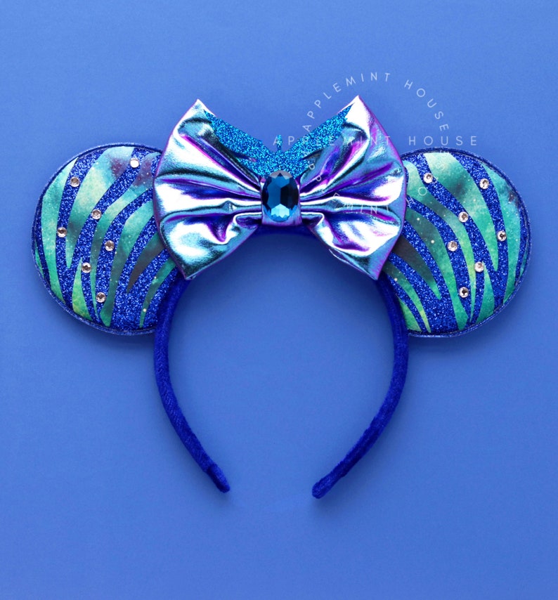Op de afbeelding: Blauw en groen glitter muis oren haarband met een holografische regenboogkleurige strik en een blauwe strass in het midden. De haarband is blauw.