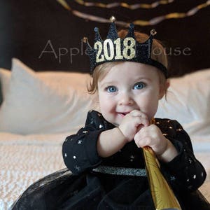 May include: A baby wearing a black and gold glitter crown with the year 2018 on it. The crown is on the baby's head and the baby is looking at the camera.