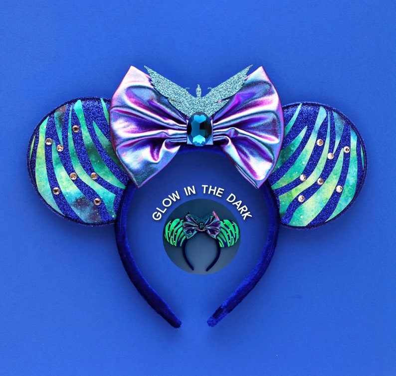 May include: Blue and green glow in the dark mouse ears with a purple iridescent bow and a blue jewel in the center. The headband is blue velvet. The text 'Glow in the Dark' is printed on a circle in the center of the image.