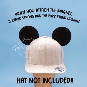 May include: A pair of black felt Mickey Mouse ears with a magnet on the back. The ears are attached to a beige baseball cap. The text "WHEN YOU ATTACH THE MAGNET, IT STAYS STRONG AND THE EARS STAND UPRIGHT" is at the top of the image. The text "HANDMAD" and "Applement House" are visible on the cap. The text "HAT NOT INCLUDED!!" is at the bottom of the image.