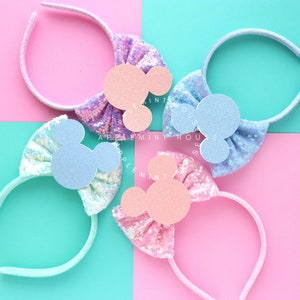 Puede incluir: Tres diademas de colores pastel con lazos de lentejuelas y orejas de Mickey Mouse de fieltro. Las diademas son azul claro, rosa y morado.
