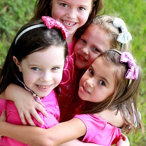 May include: Three young girls wearing pink shirts and glitter bow headbands are hugging each other. The text "You can make Glitter Bow Headband!" is at the top of the image. The image is taken outdoors with a green grassy background. The text "Amanda Anselmi Photography" is at the bottom of the image.