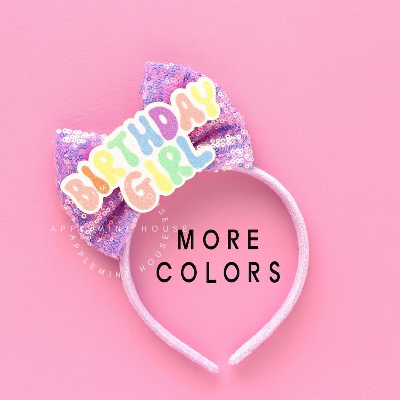 Puede incluir: Una diadema rosa con un lazo brillante y una aplicaci&oacute;n de "Birthday Girl" de colores arco&iacute;ris. La diadema est&aacute; sobre un fondo rosa con el texto "MORE COLORS" debajo.