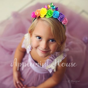 May include: A young girl wearing a purple dress and a rainbow flower headband with rhinestones. The headband is on a silver band. The girl is smiling and looking at the camera. The text "Applemint House" is visible in the image.