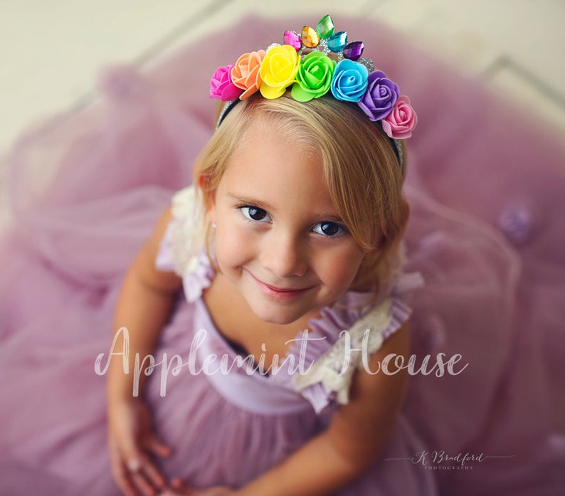 May include: A young girl wearing a purple dress and a rainbow flower headband with rhinestones. The headband is on a silver band. The girl is smiling and looking at the camera. The text "Applemint House" is visible in the image.