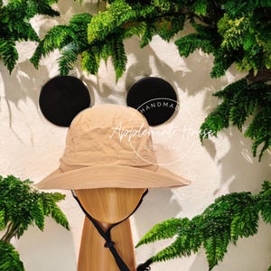 May include: A tan bucket hat with black ear flaps. The hat is on a wooden mannequin head. The hat has the text "Handmade Applemint House Est 2023" on the side.