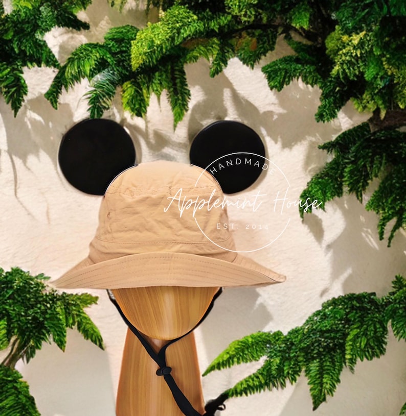 May include: A tan bucket hat with black ear flaps. The hat is on a wooden mannequin head. The hat has the text "Handmade Applemint House Est 2023" on the side.