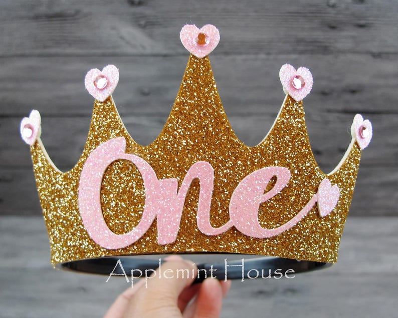 May include: Gold glitter crown with pink glitter accents and the word "One" in pink glitter. The crown is on a black headband.