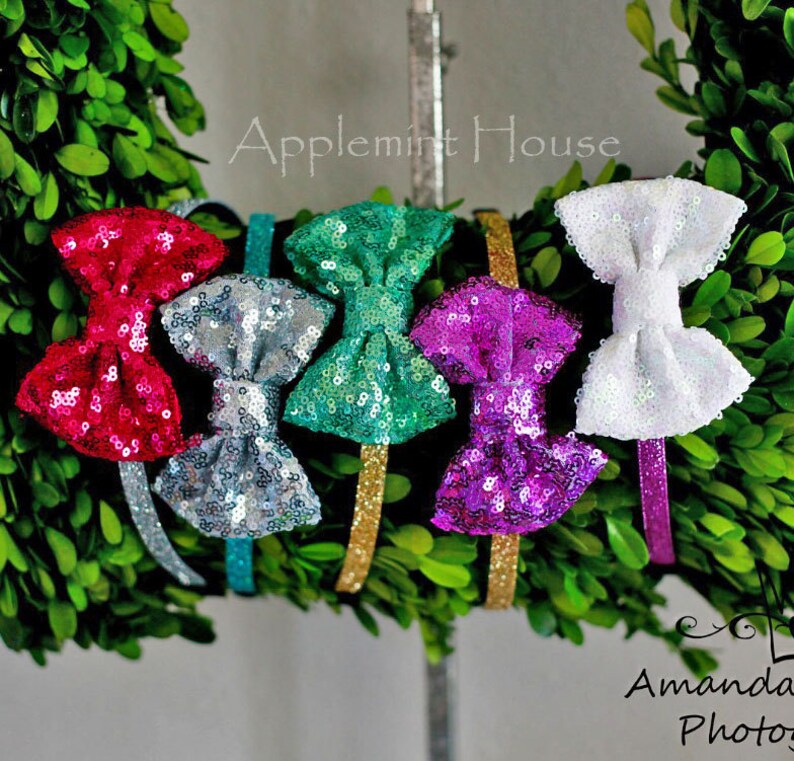 Pu&ograve; includere: Sei fasce per capelli con fiocco di paillettes scintillanti in vari colori, tra cui rosa, verde acqua, argento, viola e bianco. Le fasce sono esposte su una ghirlanda verde con il testo "Applemint House" sopra.