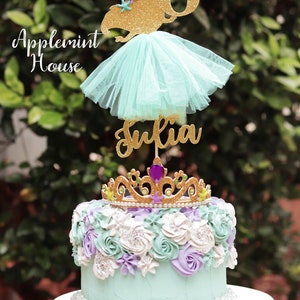 May include: A turquoise and purple frosted cake with a gold glitter crown and a gold glitter mermaid topper with a tulle skirt. The topper says "Julia" and "8".