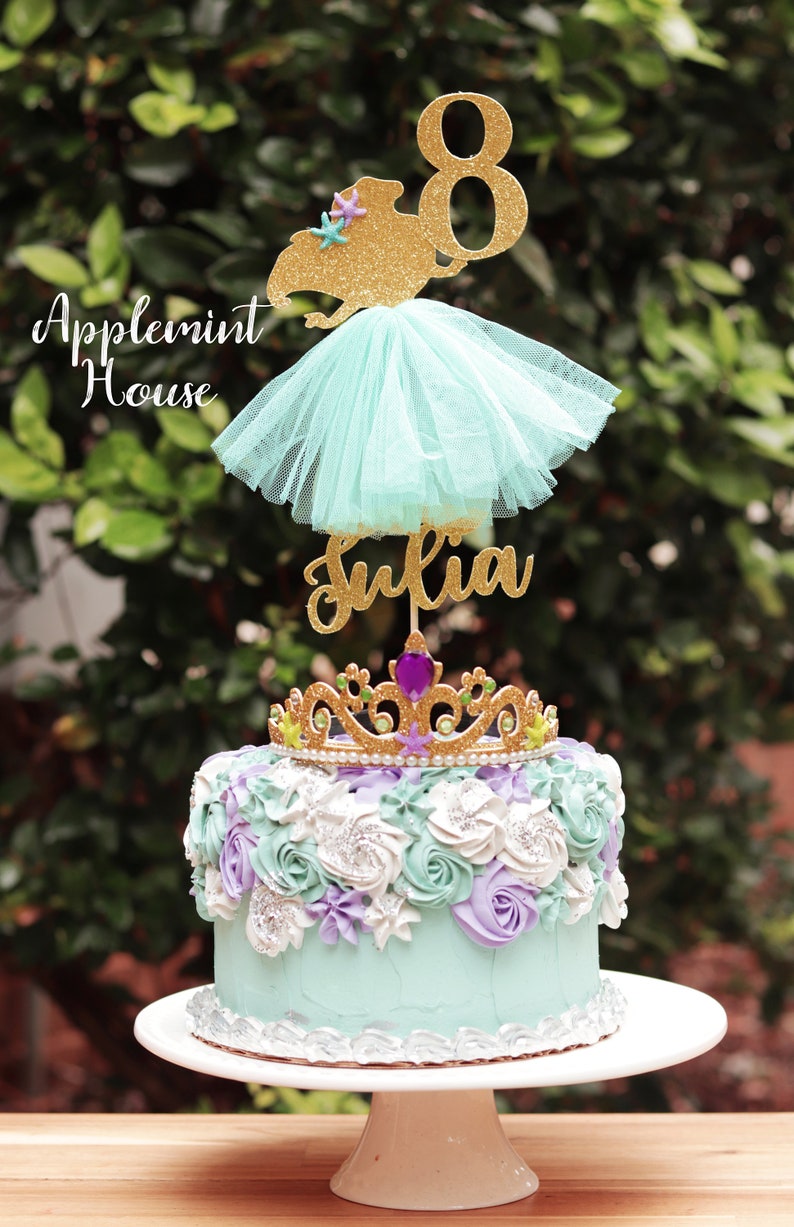 May include: A turquoise and purple frosted cake with a gold glitter crown and a gold glitter mermaid topper with a tulle skirt. The topper says "Julia" and "8".