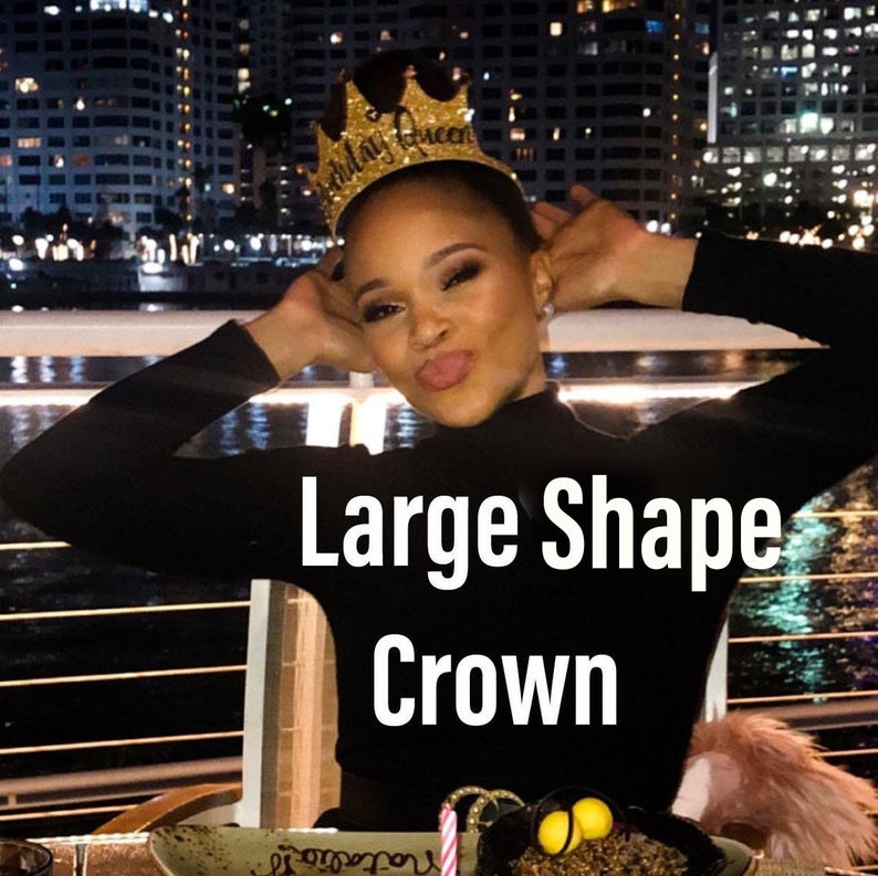 May include: A woman wearing a black shirt and a gold glitter crown that says "Birthday Queen". The crown is large and has a wide shape. The woman is smiling and looking at the camera.