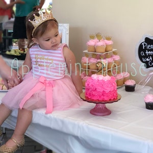 May include: A young girl wearing a pink and white striped dress with a pink tulle skirt and a gold glitter crown headband. She is sitting at a table with a pink and white frosted cake decorated with a gold number 2 and pink roses. There are cupcakes with pink frosting and gold crowns on a tiered stand behind the cake.