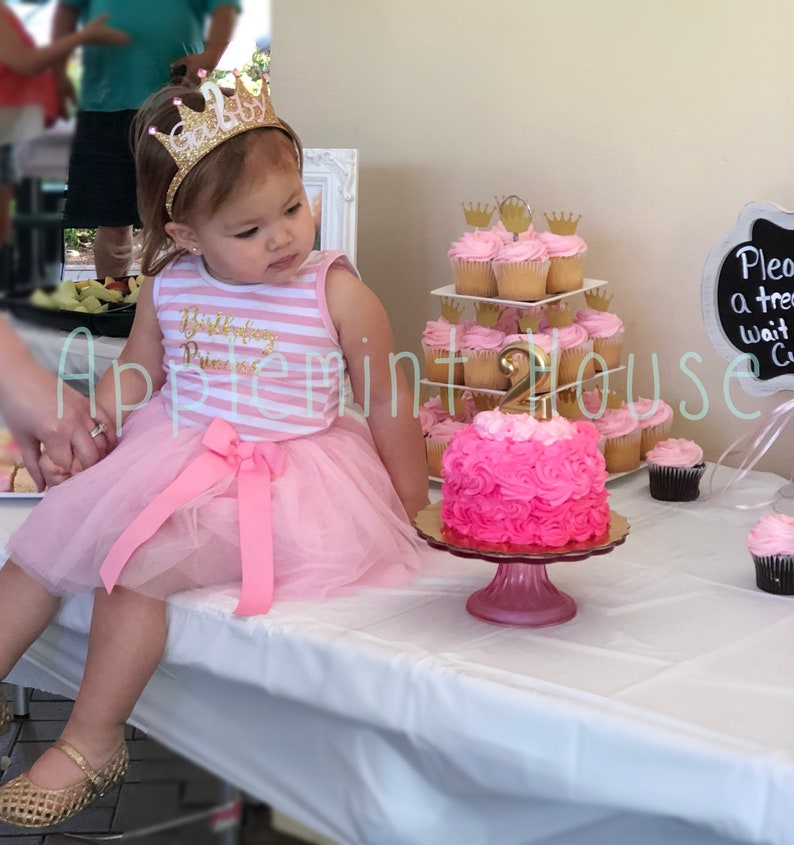 May include: A young girl wearing a pink and white striped dress with a pink tulle skirt and a gold glitter crown headband. She is sitting at a table with a pink and white frosted cake decorated with a gold number 2 and pink roses. There are cupcakes with pink frosting and gold crowns on a tiered stand behind the cake.