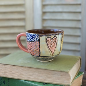 Pode incluir: Uma caneca de cerâmica artesanal com um design de patchwork. A caneca apresenta uma alça rosa, um interior castanho e um desenho de coração na frente. O exterior tem uma mistura de padrões e cores, incluindo azul, rosa e amarelo. Está sobre uma pilha de livros antigos.