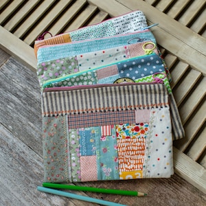 May include: A stack of colorful, patchwork fabric pencil cases with zipper closures. The cases feature various patterns, including floral, polka dots, and stripes. Two pencils, one green and one blue, are lying on the wooden surface.