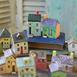 Puede incluir: Una colección de casas en miniatura de cerámica colorida. Cada casa presenta detalles únicos como diferentes estilos de techo, colores de puertas y diseños de ventanas. Las casas están dispuestas sobre una superficie de madera, creando una escena de pueblo encantadora.