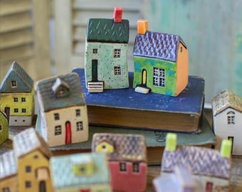 May include: A collection of colorful ceramic miniature houses. Each house features unique details like different roof styles, door colors, and window arrangements. The houses are arranged on a wooden surface, creating a charming village scene.
