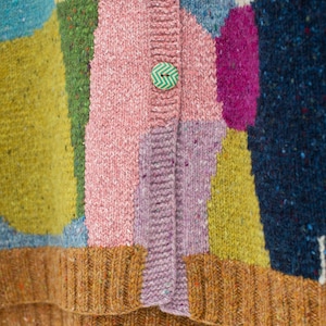 Może przedstawiać: Zbliżenie kolorowego kardiganu w stylu patchwork. Sweter ma r&oacute;żne kolory, w tym r&oacute;żowy, niebieski, zielony, ż&oacute;łty i fioletowy. Ma dwa ozdobne guziki i prążkowany brązowy d&oacute;ł.