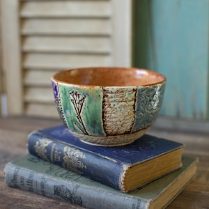Può includere: Una ciotola decorativa in ceramica con un design patchwork nei toni del verde, blu e marrone, con motivi floreali e di scrittura. La ciotola è appoggiata su due libri antichi con scritte dorate, creando un'estetica vintage.