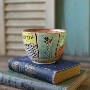 May include: A handcrafted ceramic bowl with a patchwork design. The bowl features sections of yellow, blue, green, and orange, with the word "verace" on the yellow section. A floral design is on the blue section. The bowl is sitting on top of two antique books.