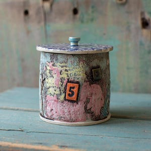 May include: A cylindrical ceramic jar with a lid. The jar is painted with a textured, colorful design in shades of pink, yellow, and blue. The lid is decorated with a blue and white pattern. The number "5" is displayed on the front.