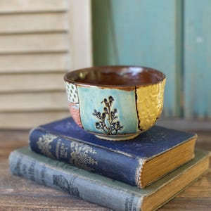 Könnte beinhalten: Handgefertigte Keramikschale mit braunem Innenraum und Patchwork-Außenbereich. Die Außenseite hat Abschnitte in Blau, Gelb und gemusterten Designs. Die Schale liegt auf zwei antiken Büchern mit goldener Schrift.