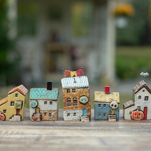 May include: A set of five miniature ceramic houses featuring Halloween-themed details. The houses include a yellow building with a pumpkin, a white house with a green roof and skull, a tall brown building with bunting, a blue house with a skull, and a white house with a pumpkin.