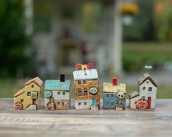 May include: A set of five miniature ceramic houses featuring Halloween-themed details. The houses include a yellow building with a pumpkin, a white house with a green roof and skull, a tall brown building with bunting, a blue house with a skull, and a white house with a pumpkin.