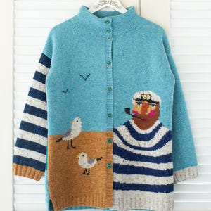 Puede incluir: Cárdigan de punto azul claro con temática náutica. El suéter presenta un diseño de marinero, gaviotas y mangas a rayas. El cárdigan tiene cuello alto y cierre de botones. Las medidas no están disponibles.